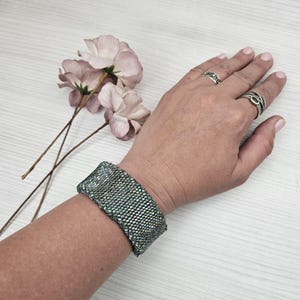 May include: A wide, beaded bracelet in shades of green and gray is worn on a wrist. The bracelet has a textured appearance. Two silver rings are on the fingers. Pink flowers are in the background.