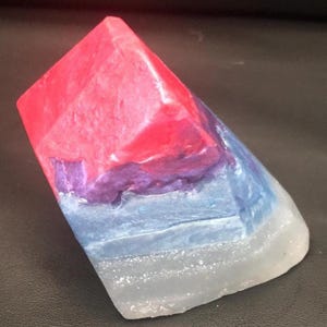 May include: A multi-colored, geometric soap bar. The top is a vibrant red, transitioning to purple, blue, and a translucent white base. The soap has a faceted, angular design, resembling a crystal or gemstone. The soap is a unique decorative item.