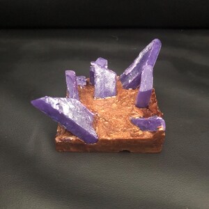 May include: A small, square sculpture featuring a textured, brown base with several purple crystal formations. The crystals vary in size and shape, with some appearing sharp and pointed.