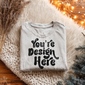 Op de afbeelding: Een lichtgrijs t-shirt met de tekst "You're Design Here" in zwart, weergegeven op een witte, zachte ondergrond. Het shirt is netjes opgevouwen en toont het ontwerp. De afbeelding heeft een gezellige, uitnodigende uitstraling.