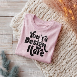 May include: A light pink t-shirt with the text "You're Design Here" in a black, bold, bubble font. The shirt is folded and resting on a cream-colored knitted blanket. The background includes a wooden surface and decorative elements.