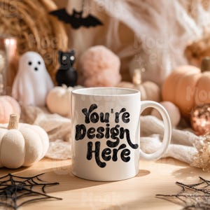 Pode incluir: Uma caneca de cerâmica branca com o texto "You're Design Here" em preto. A caneca está sobre uma superfície de madeira, rodeada por decorações de Halloween, incluindo abóboras, uma teia de aranha e um morcego.