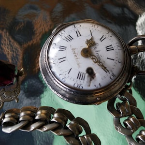 Könnte beinhalten: Antike Taschenuhr mit weißem Zifferblatt, römischen Ziffern und verzierten Zeigern. Die Uhr ist an einer silbernen Kette mit einem dekorativen Verschluss befestigt, der einen dunkelroten Edelstein hält. Das Zifferblatt der Uhr zeigt "Herrenkohl".