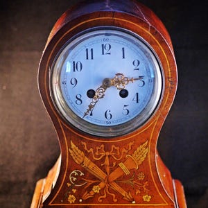Puede incluir: Un reloj antiguo de madera con una esfera azul claro y manecillas doradas. El marco de madera del reloj está decorado con diseños incrustados de trigo y motivos florales. El reloj es un objeto decorativo.