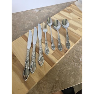 Peut inclure: Un ensemble de couverts en acier inoxydable, comprenant des couteaux, des fourchettes et des cuillères, disposés sur une planche à découper en bois. Les couverts présentent des détails floraux ornés sur les manches. L'ensemble comprend six couteaux, une fourchette et quatre cuillères.