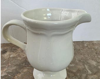 May include: A white ceramic pitcher with a handle and a spout. The pitcher has a bulbous body and sits on a pedestal base. The handle is curved, and the spout is slightly flared. The pitcher is likely used for serving liquids.