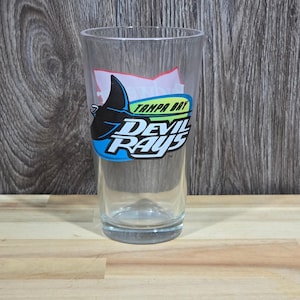 Peut inclure: Un verre transparent avec le logo des Tampa Bay Devil Rays. Le logo présente une raie manta noire et bleue et les mots "Tampa Bay Devil Rays" en blanc et bleu. Le verre est cylindrique et vide.