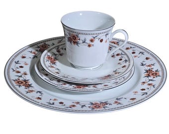 May include: A white porcelain teacup, saucer, and two plates set. The set features a floral pattern in shades of orange, brown, and blue. The edges of the plates and cup are trimmed with a dark gray border.