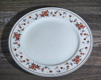 May include: White oval serving platter with a floral design. The rim features a delicate pattern with orange and blue flowers and green leaves. The plate has a silver trim and is set against a wooden surface.