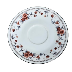 May include: White saucer with a floral design. The saucer features a central recessed area and is decorated with a border of pink and brown flowers with blue leaves. A thin black line borders the edge.