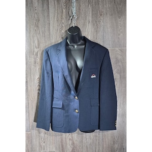 May include: A navy blue blazer with gold buttons and two front pockets. The blazer has a small logo on the left chest. The blazer is displayed on a black mannequin against a wooden background.