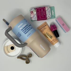 May include: A beige Owala insulated tumbler with a gray handle and straw, next to a manicure set, hand cream, nail polish, and a nail file. The tumbler has a blue label with text. A hair clip and tumbler lid are also present.