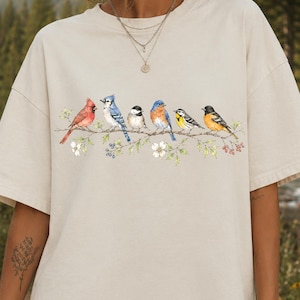 May include: A beige short-sleeve t-shirt featuring a graphic print of six colorful birds perched on a flowering branch. The birds include a red cardinal, a blue jay, a black and white chickadee, a bluebird, a yellow warbler, and an oriole.