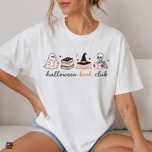 Puede incluir: Una camiseta blanca con un estampado gr&aacute;fico que muestra un fantasma, una pila de libros, un sombrero de bruja sobre un libro abierto y un esqueleto leyendo. El texto "halloween book club" est&aacute; impreso en letras negras y naranjas.