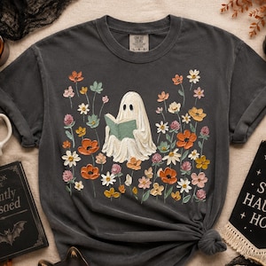 May include: A charcoal gray short-sleeve t-shirt featuring a central graphic of a white ghost reading a green book, surrounded by a colorful array of painted-style flowers in shades of orange, yellow, pink, and white, with a Comfort Colors label.