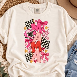 May include: A cream-coloured short-sleeve t-shirt featuring a graphic print of the word MAMA in pink and red letters. The design includes a pink motocross helmet, black and white checkered racing flags, pink bows, yellow stars, and lightning bolts.