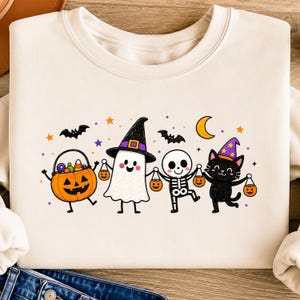 May include: A cream-coloured crewneck sweatshirt featuring a colourful Halloween graphic of a jack-o'-lantern, a ghost wearing a witch hat, a skeleton, and a black cat wearing a purple hat, all holding small orange pumpkin-shaped candy buckets.