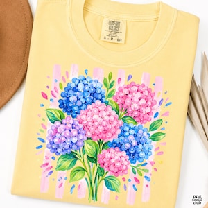 Op de afbeelding: Een lichtgeel t-shirt met een grafische print van een boeket hortensia's met blauwe, roze en paarse bloemen en groene bladeren. Het ontwerp is geplaatst tegen verticale lichtroze strepen. Het binnenlabel vermeldt de tekst Comfort Colors.