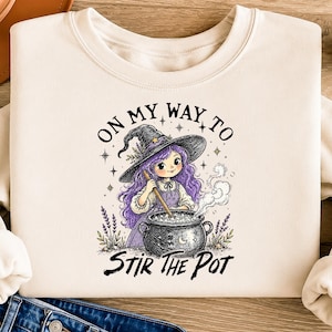 May include: A cream-colored sweatshirt featuring a graphic illustration of a character with purple hair wearing a witch hat, stirring a steaming black cauldron. The text reads ON MY WAY TO STIR THE POT, surrounded by small stars and lavender sprigs.