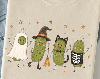 Könnte beinhalten: Ein cremefarbenes T-Shirt mit einer Grafik von vier grünen Gewürzgurken in Halloween-Kostümen: ein Geist, eine Hexe mit Besen, eine schwarze Katze und ein Skelett. Das Design enthält kleine orange, grüne und braune Sterne im Hintergrund.