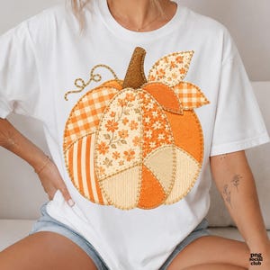 May include: A white short-sleeve t-shirt featuring a large central graphic of a pumpkin constructed from various fabric patches. The pumpkin design includes orange and white gingham, floral patterns, and solid orange sections with visible stitching.