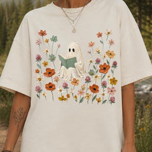 May include: A cream-colored short-sleeve t-shirt featuring a central graphic of a white ghost reading a green book, surrounded by a field of textured, colorful wildflowers in shades of orange, pink, yellow, and green, rendered in an impasto style.