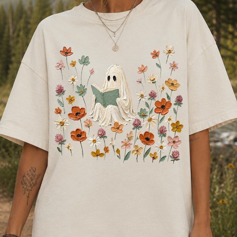 May include: A cream-colored short-sleeve t-shirt featuring a central graphic of a white ghost reading a green book, surrounded by a field of textured, colorful wildflowers in shades of orange, pink, yellow, and green, rendered in an impasto style.