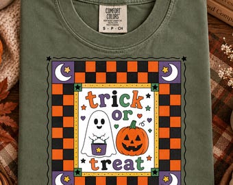 Op de afbeelding: Een olijfgroen t-shirt met ronde hals en een Comfort Colors-label. De afbeelding heeft een zwart-oranje geblokte rand met witte maansikkels en sterren. In het midden staat een geest met een paarse emmer en een pompoen afgebeeld.