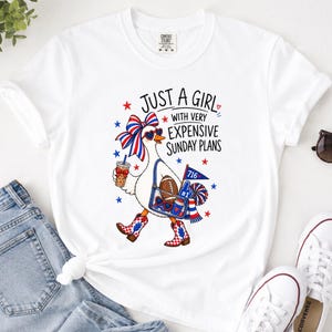 May include: A white short-sleeve t-shirt featuring a graphic of a goose wearing a red, white, and blue bow, heart-shaped sunglasses, and patterned boots. The goose carries a bag with a football, a pennant labeled 716, and a drink. Text reads: JUST A GIRL WITH VERY EXPENSIVE SUNDAY PLANS.