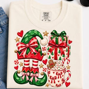 Peut inclure: Un t-shirt couleur crème avec une impression graphique représentant des jambes d'elfe en bas rayés et chaussures vertes, des cadeaux empilés avec des nœuds rouges, et une tasse garnie de crème fouettée, un biscuit pain d'épices et un sucre d'orge.