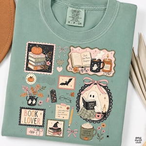 K&ouml;nnte beinhalten: Ein salbeigr&uuml;nes T-Shirt mit einem Grafikdruck herbstlicher Illustrationen, darunter B&uuml;cherstapel, ein lesendes Gespenst, K&uuml;rbisse, eine Tasse mit Gespenst, eine Kerze, eine Fledermaus, eine Brille, ein Bleistift und der Schriftzug Book Lover.