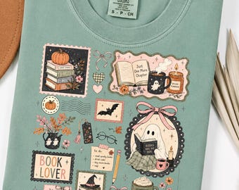 May include: A sage green t-shirt featuring a graphic print of autumn-themed illustrations including stacks of books, a ghost reading a book, pumpkins, a mug with a ghost, a candle, a bat, glasses, a pencil, and text reading Book Lover.