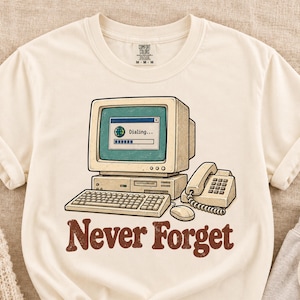 May include: A cream-colored t-shirt featuring a graphic of a vintage beige computer monitor, keyboard, mouse, and landline telephone. The screen displays a loading window with the text Dialing. Below the illustration, the text Never Forget is printed in a brown, bold, serif font.