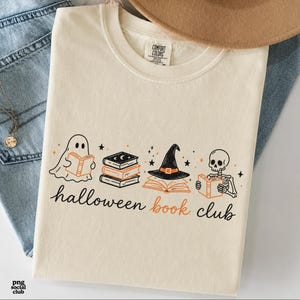 Puede incluir: Una camiseta de cuello redondo color crema con un estampado gr&aacute;fico de un fantasma, una pila de libros, un sombrero de bruja sobre un libro abierto y un esqueleto, todos leyendo. El texto bajo las ilustraciones dice "halloween book club".