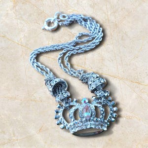 May include: Silver necklace with a crown pendant. The crown is decorated with clear and iridescent rhinestones, and a central oval stone. The necklace has a multi-strand chain and a lobster clasp.
