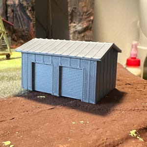May include: A miniature gray model of a two-car garage with a corrugated metal roof. The garage has two doors and is set on a brown, textured surface, with a backdrop of a rocky mountain and a small swing set.