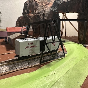 May include: A model train scene featuring an Erie Lackawanna boxcar on tracks, passing under a black gantry. The scene includes miniature buildings, a rock formation, and a green painted road. The boxcar is white with red lettering.