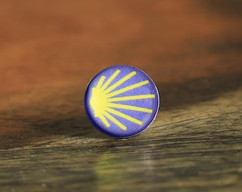 May include: A small, circular pin featuring a vibrant blue background with a yellow scallop shell graphic. The design displays radiating lines originating from the left side of the shell, creating a distinct sunburst pattern on the round surface.