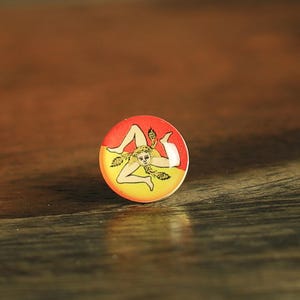 May include: A round button with a red and yellow background featuring a Trinacria symbol. The Trinacria symbol is a figure with three legs, a face, and wheat. The button is on a wooden surface.