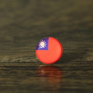 May include: A small, round pin featuring the flag of Taiwan. The pin has a red background with a blue square in the upper left corner, containing a white sun with rays. The pin is set against a dark wood grain surface.