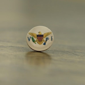 May include: A round, white pin featuring the emblem of the U.S. Virgin Islands. The emblem includes a gold eagle with outstretched wings, a red and blue shield, and the letters "V" and "I".
