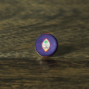May include: A round button with a purple background and the word "GUAM" in white. The button features a palm tree and a diamond shape in the center. The button has a red border and a gold metal edge.