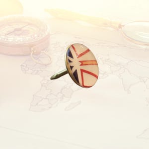 May include: An oval-shaped push pin with a Union Jack design in red, white, and blue. The pin has a silver-colored metal base and a long, pointed pin. The background includes a map, compass, and magnifying glass.