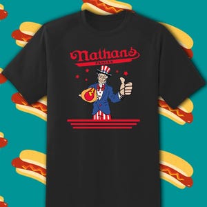 May include: Black t-shirt featuring the text "Nathan's Famous" in red script above an illustration of Uncle Sam holding a hot dog and giving a thumbs up. The design includes red stars and stripes.