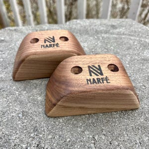 May include: Two wooden climbing holds with a curved design. Each hold has two drilled holes and the word "NARFÉ" printed below a stylized "N" logo in black. The holds are made of a dark wood and sit on a grey surface.