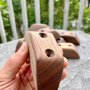 Könnte beinhalten: Eine Sammlung von Holz-Klettergriffen, jeweils mit zwei gebohrten Löchern. Die Griffe haben eine satte braune Farbe und eine glatte, abgerundete Form. Mehrere Griffe sind sichtbar, was auf ein Set für eine Kletterwand hindeutet.