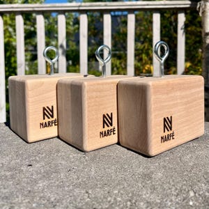 May include: Three light-colored wooden blocks with a silver metal loop on top. Each block has the word "NARFÉ" printed below a stylized "N" logo. The blocks are sitting on a gray surface, with a white fence and greenery in the background.