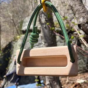 May include: A wooden climbing hold with a rectangular design and a long, recessed grip. It is suspended by a green and blue rope attached to a yellow metal ring. The hold is made of light-colored wood.