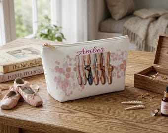 May include: A white cosmetic bag with a gold zipper, featuring a ballet-themed design. The design includes illustrations of pointe shoes and the name "Amber" in cursive. Ballet slippers and books are also visible.