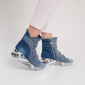Può includere: Stivaletti in denim blu con zeppa. Gli stivali sono impreziositi da piccoli strass scintillanti. La zeppa presenta un design con stampa di giornale in bianco e nero. Una cerniera e una fibbia rifiniscono il lato dello stivale.